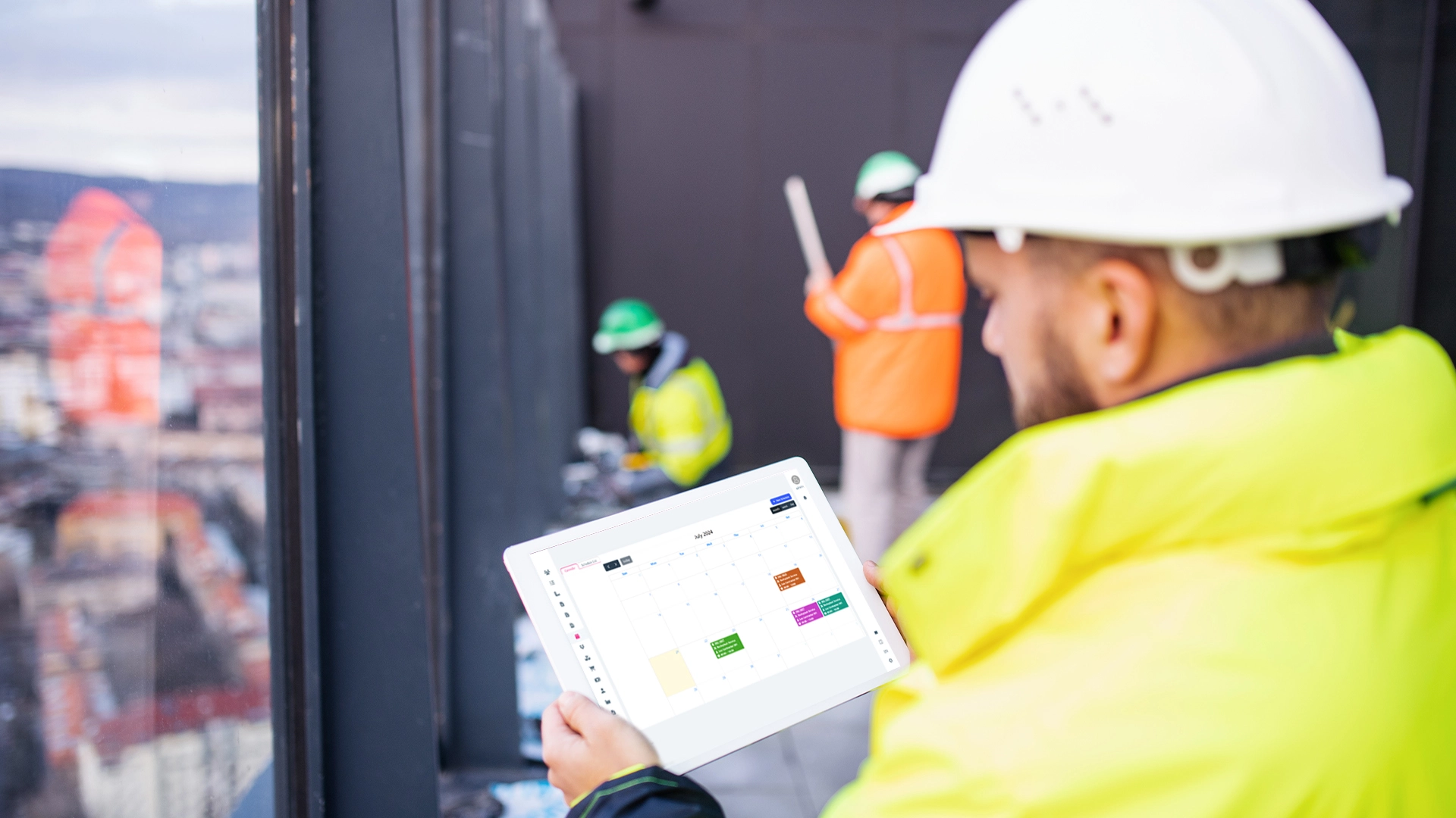 A construction worker in a hard hat and high-visibility jacket using a tablet with scheduling screen of homearize scheduling software , while other workers in safety gear operate in the background on a construction site.