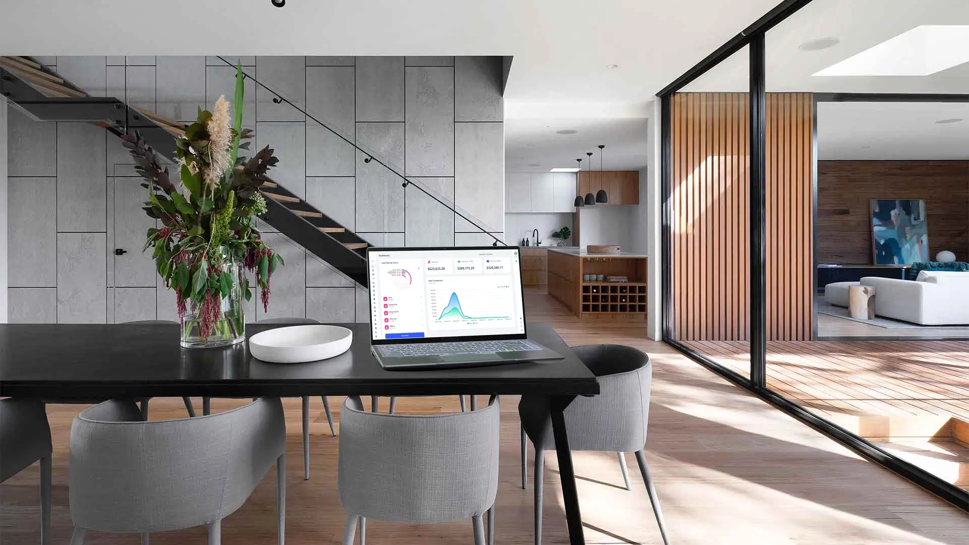 A modern home interior with a dining table featuring a laptop displaying a homearize dashboard, surrounded by elegant gray chairs and a large vase of flowers, with a wooden kitchen and living area in the background.